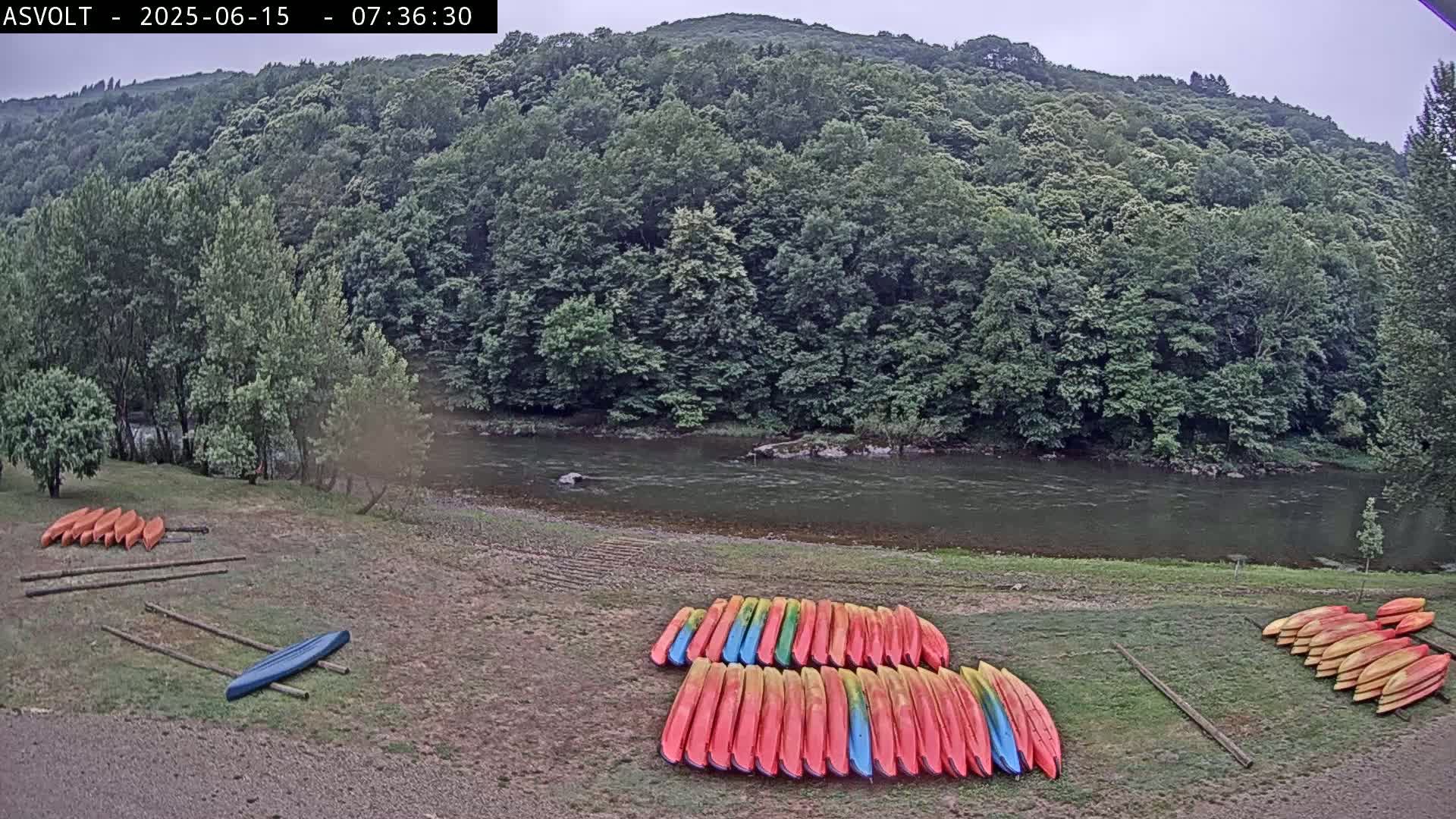 A calm river flows past a wooded hillside, where numerous kayaks are stacked on the riverbank under an overcast sky.