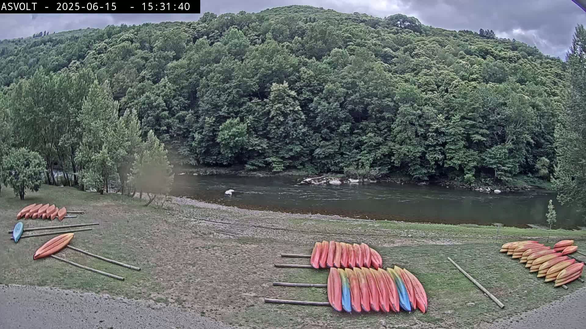 Several kayaks are stacked on the grassy bank of a calm river under a cloudy sky, next to a lush, green hillside.