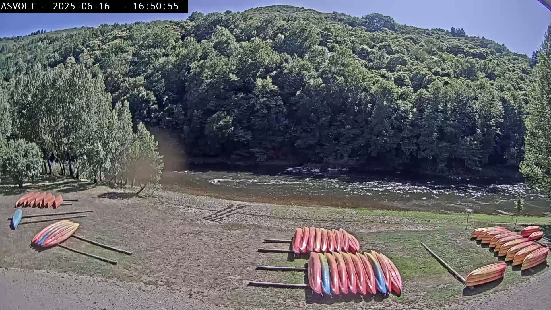 Under a sunny sky, several kayaks are stacked on the sandy bank of a river flowing beside a lush green forest.