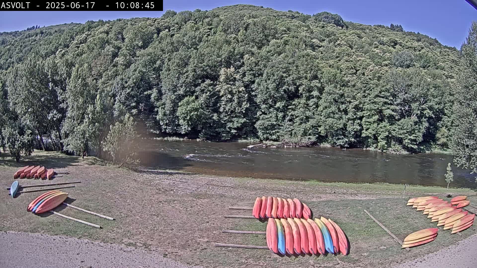 Several kayaks are stacked on the grassy bank of a river under a sunny sky, with a lush green forest visible in the background.