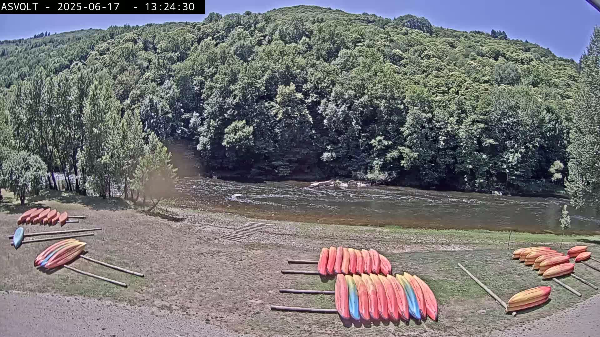 Several rows of kayaks are resting on the shore of a river, under a sunny sky, backed by a lush green forest.