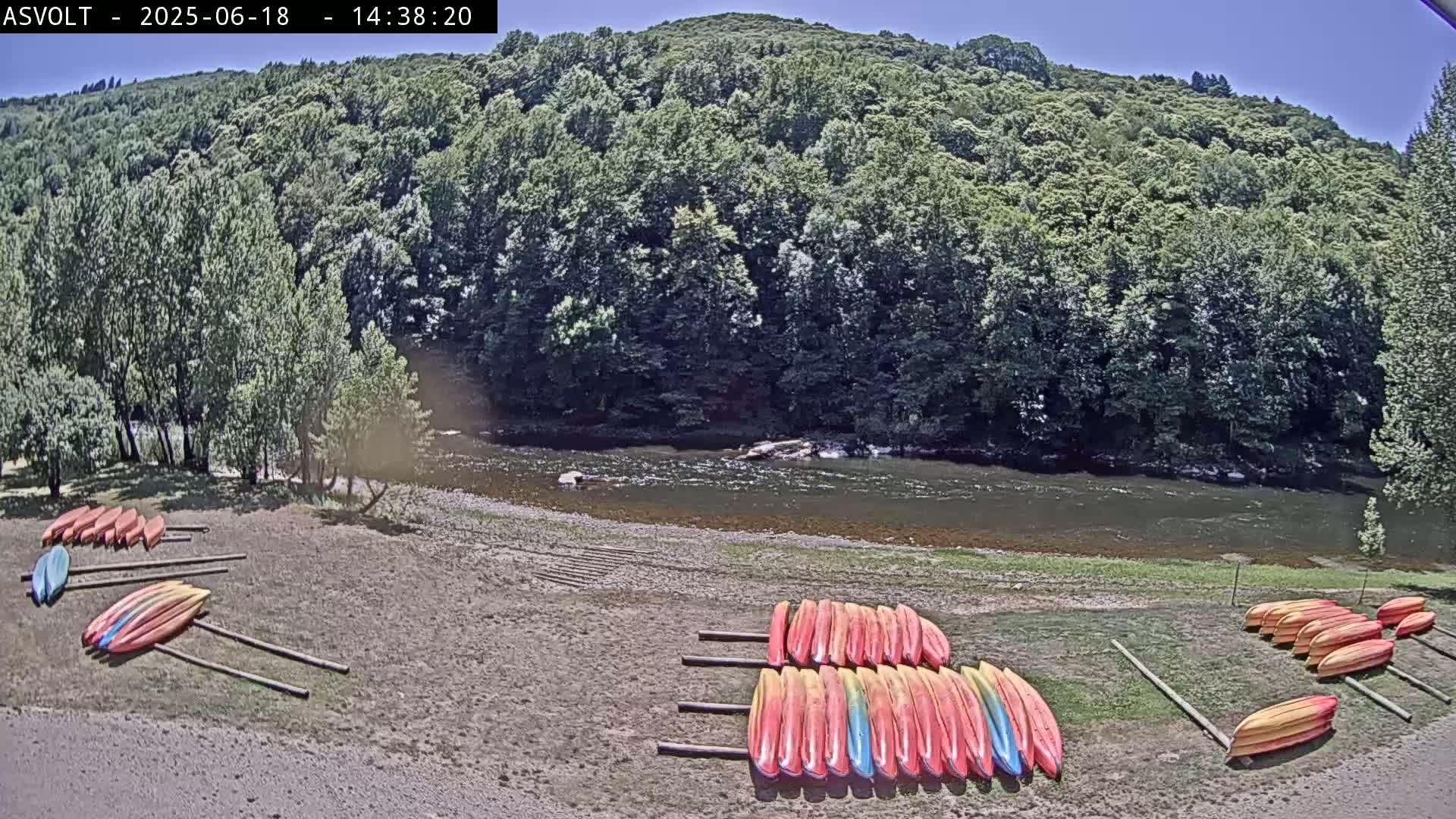 Several groups of kayaks are stacked on the sandy bank of a calm river, under a sunny sky, with a lush green forest rising up behind it.