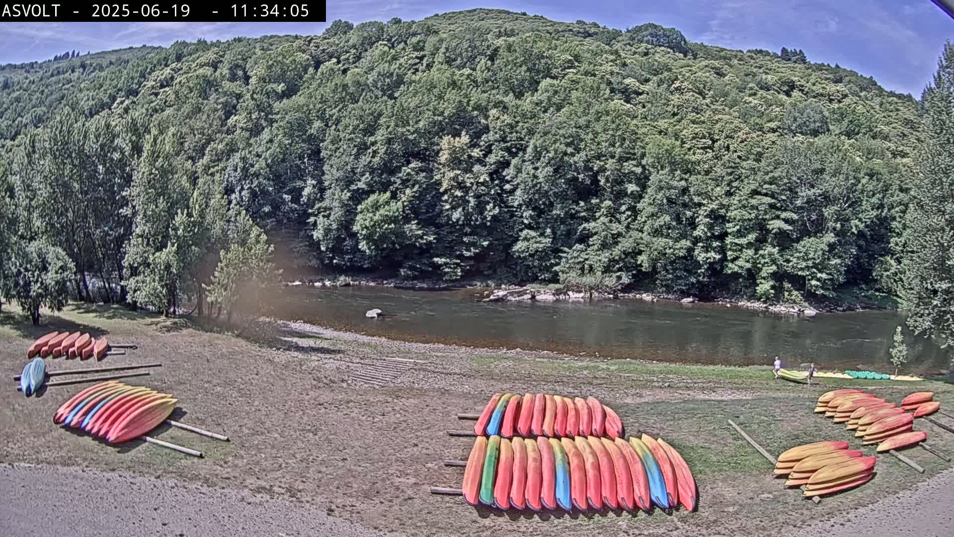 Several stacks of colorful kayaks are situated on a sandy riverbank next to a calm river flowing through a lush green forest under a clear blue sky.