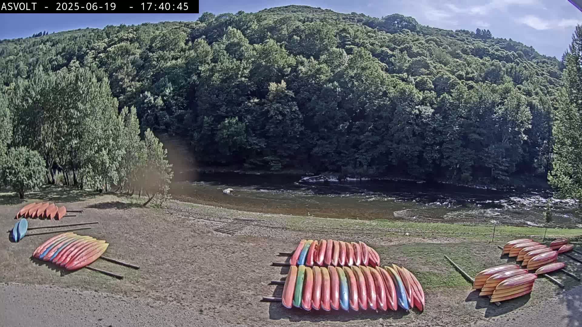Several stacks of kayaks are resting on the shore of a calm river, backed by a lush green forest under a mostly clear sky.