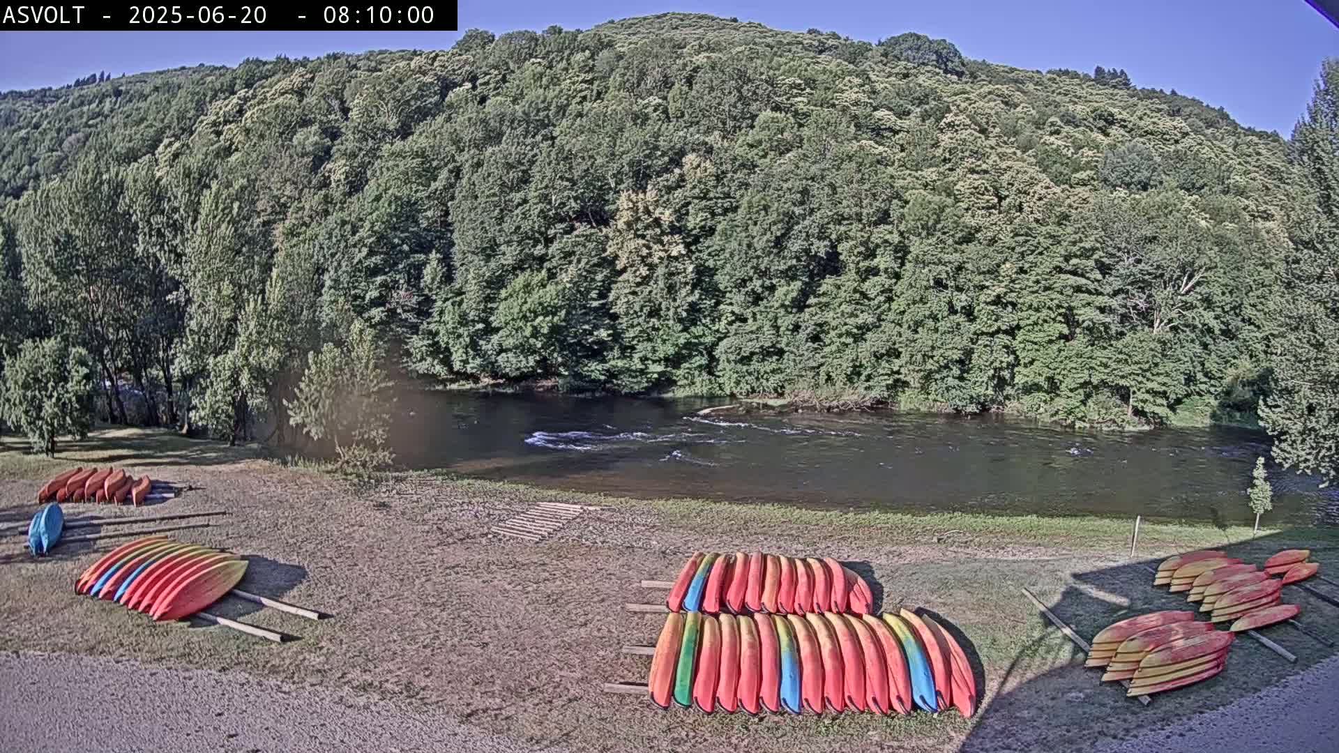 On a sunny day, several colorful kayaks are stacked on the shore of a calm river, bordered by lush green trees and hills.