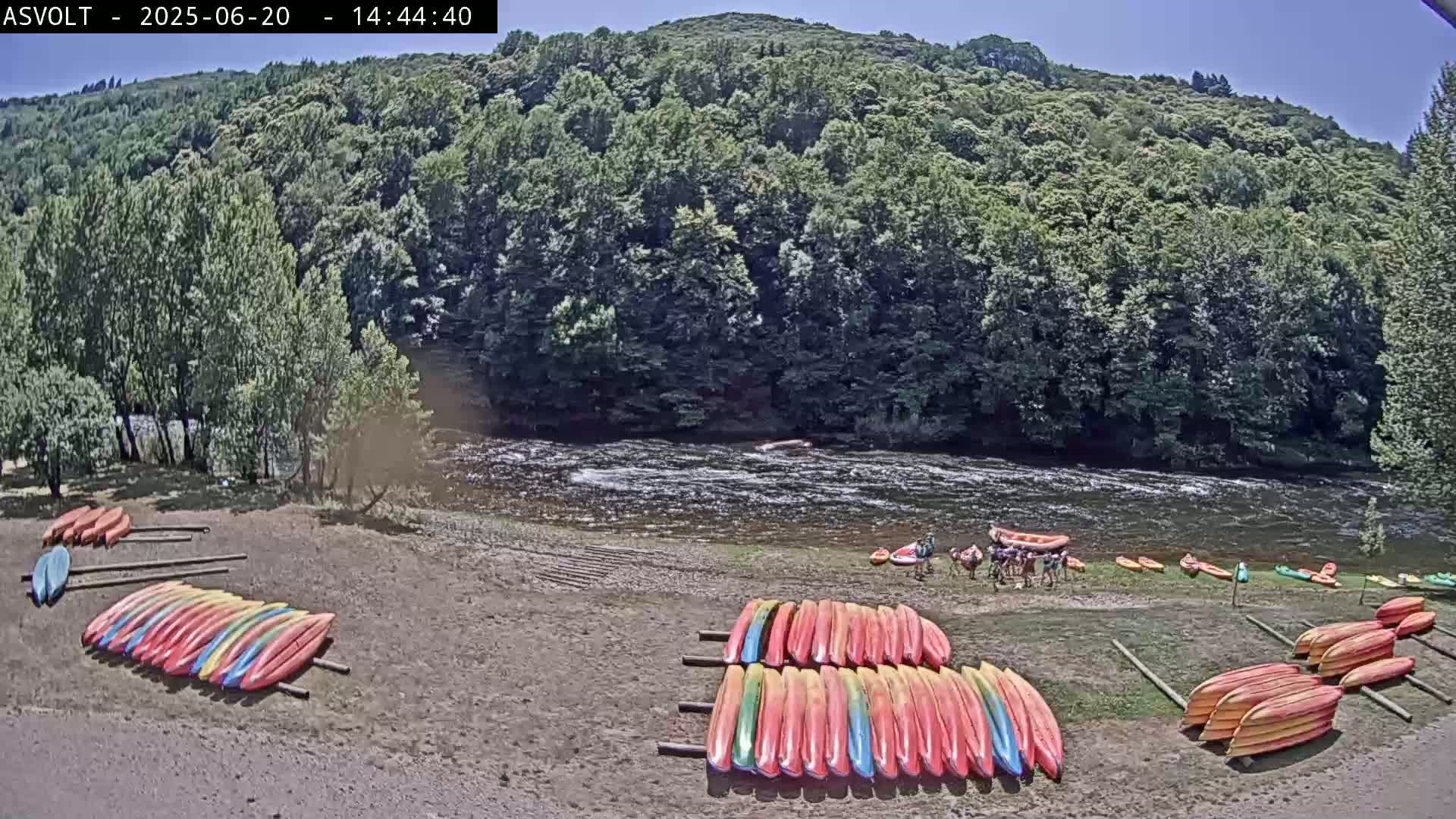 A sunny day shows a sandy riverbank with colorful kayaks stacked neatly, a few people near some kayaks on the water, and a forested hillside in the background.