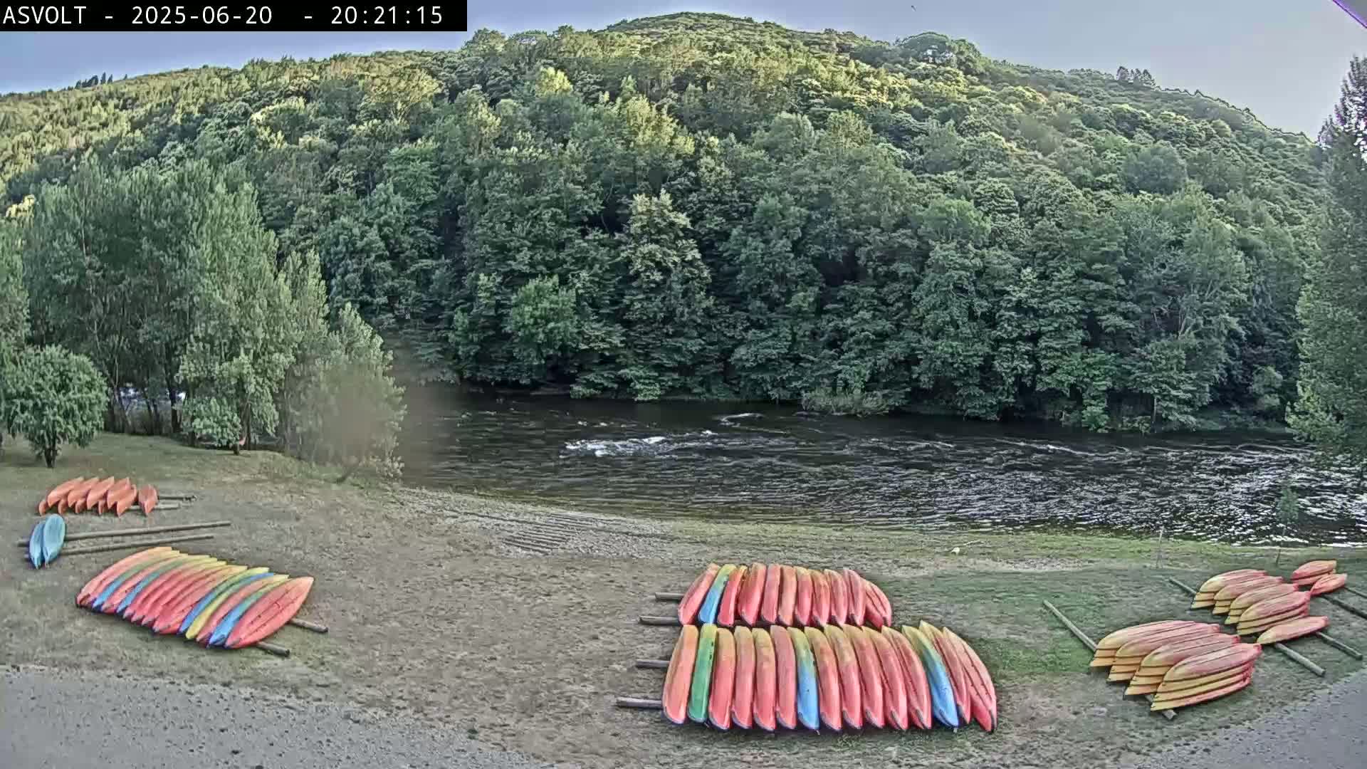 Several stacks of colorful kayaks are resting on a sandy riverbank beside a calm river under a partly cloudy sky, with lush green forest rising up beyond the river.