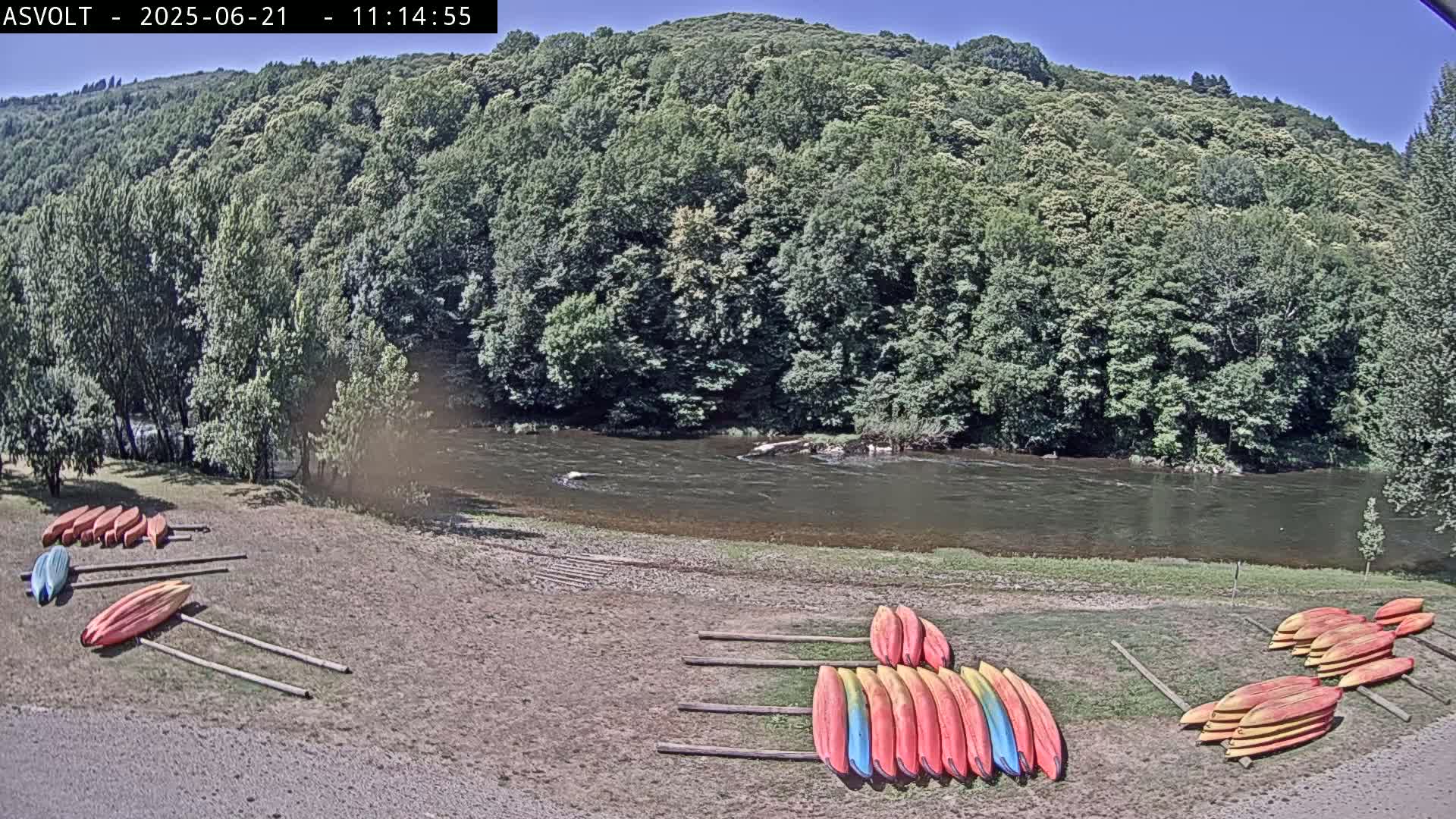 Kayaks are stacked on racks near a calm river under a clear blue sky, with lush green trees rising up a hillside in the background.