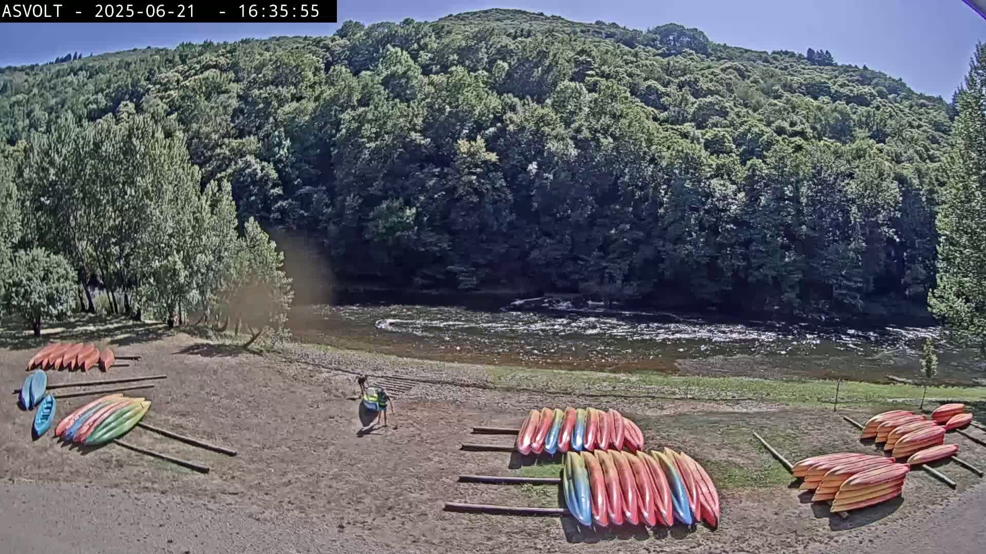 On a sunny day, colorful kayaks are stacked on the sandy bank of a river flowing past a lush green forest.