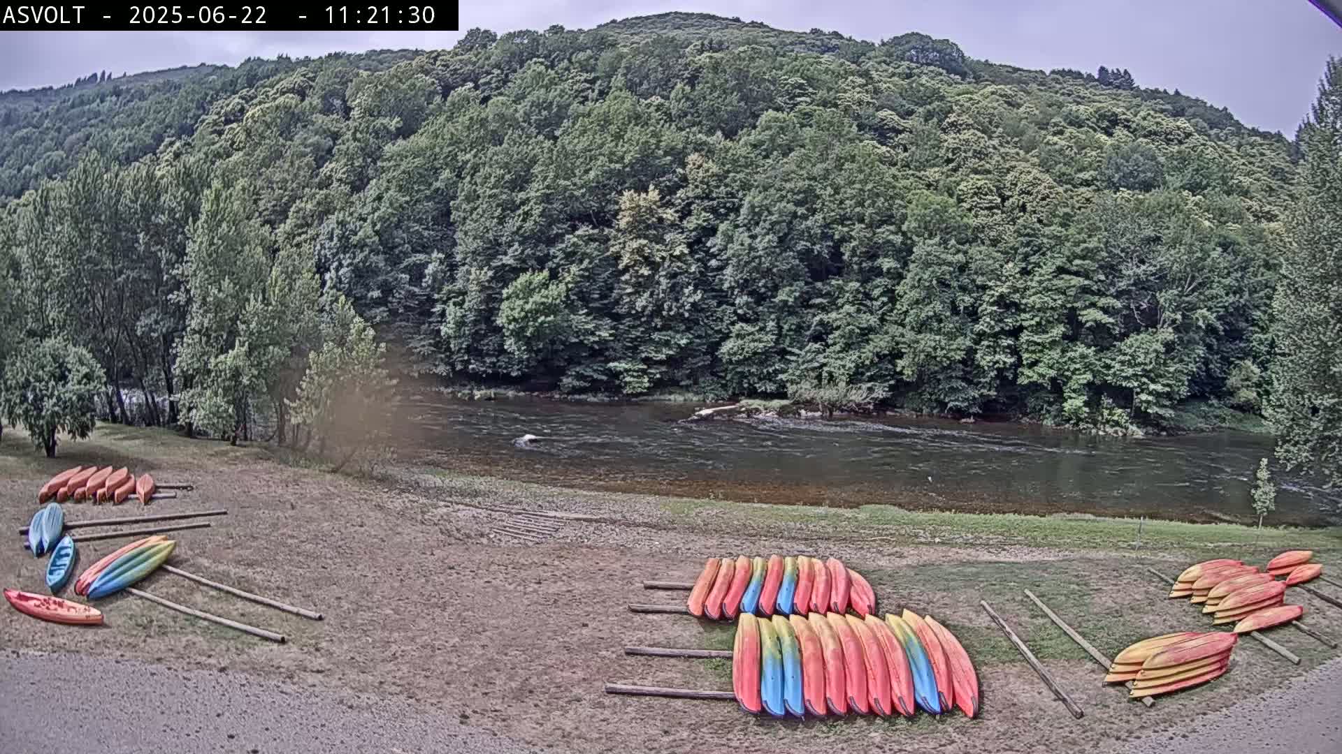 Kayaks are stacked on the shore of a calm river, next to a lush green forest under an overcast sky.