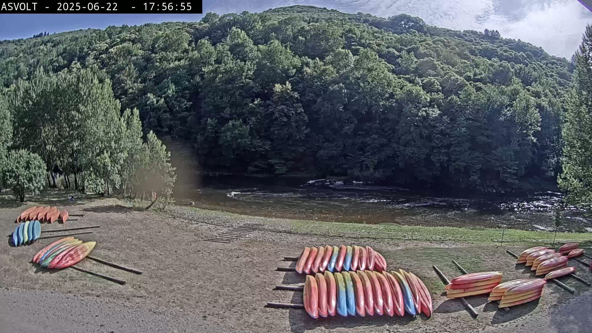 Several stacks of colorful kayaks sit on a sandy shore next to a calm river, with lush green hills rising in the background under a partly cloudy sky.