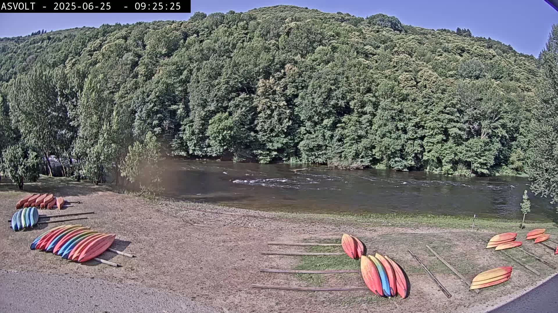 A river flows under a sunny sky past a lush green hillside where colorful kayaks are lined up on the shore.