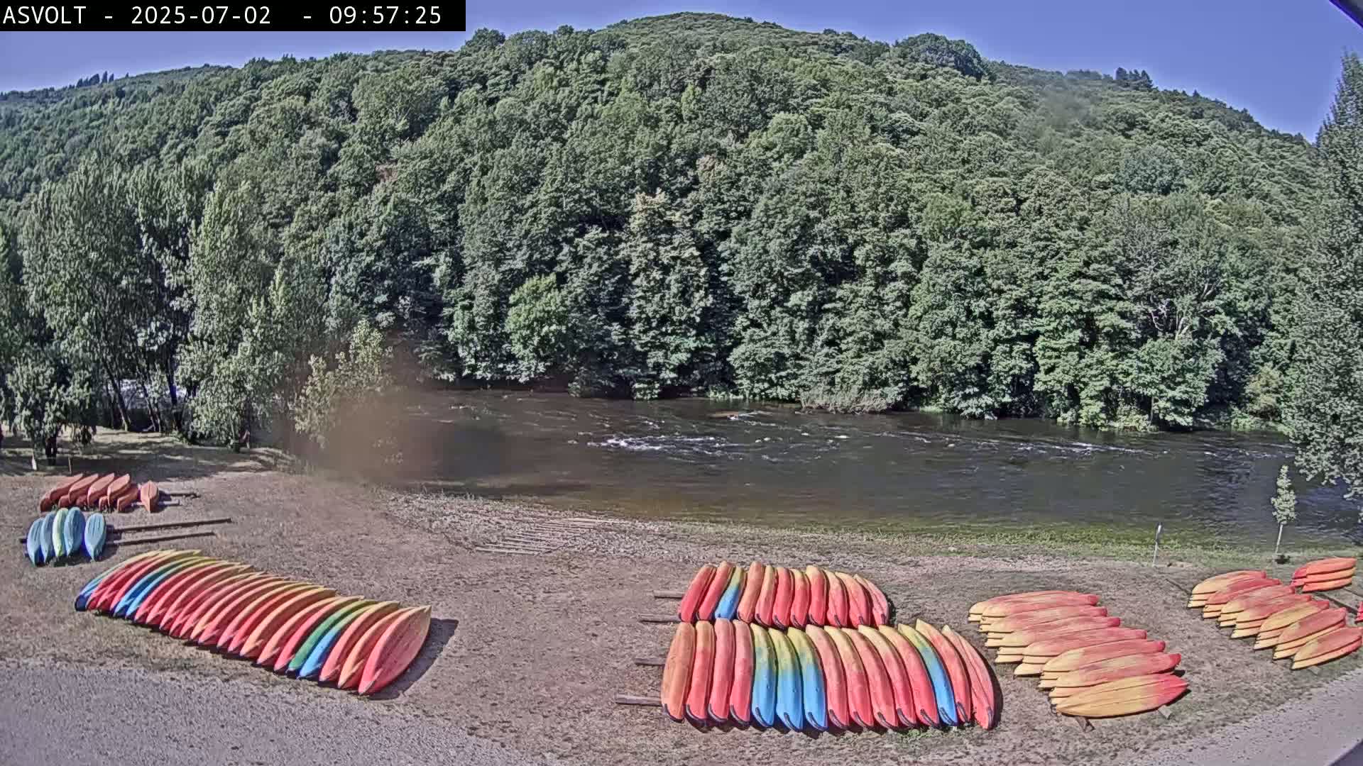 Numerous kayaks are stacked on a sandy shore next to a river flowing under a sunny sky, backed by a lush, green hillside.