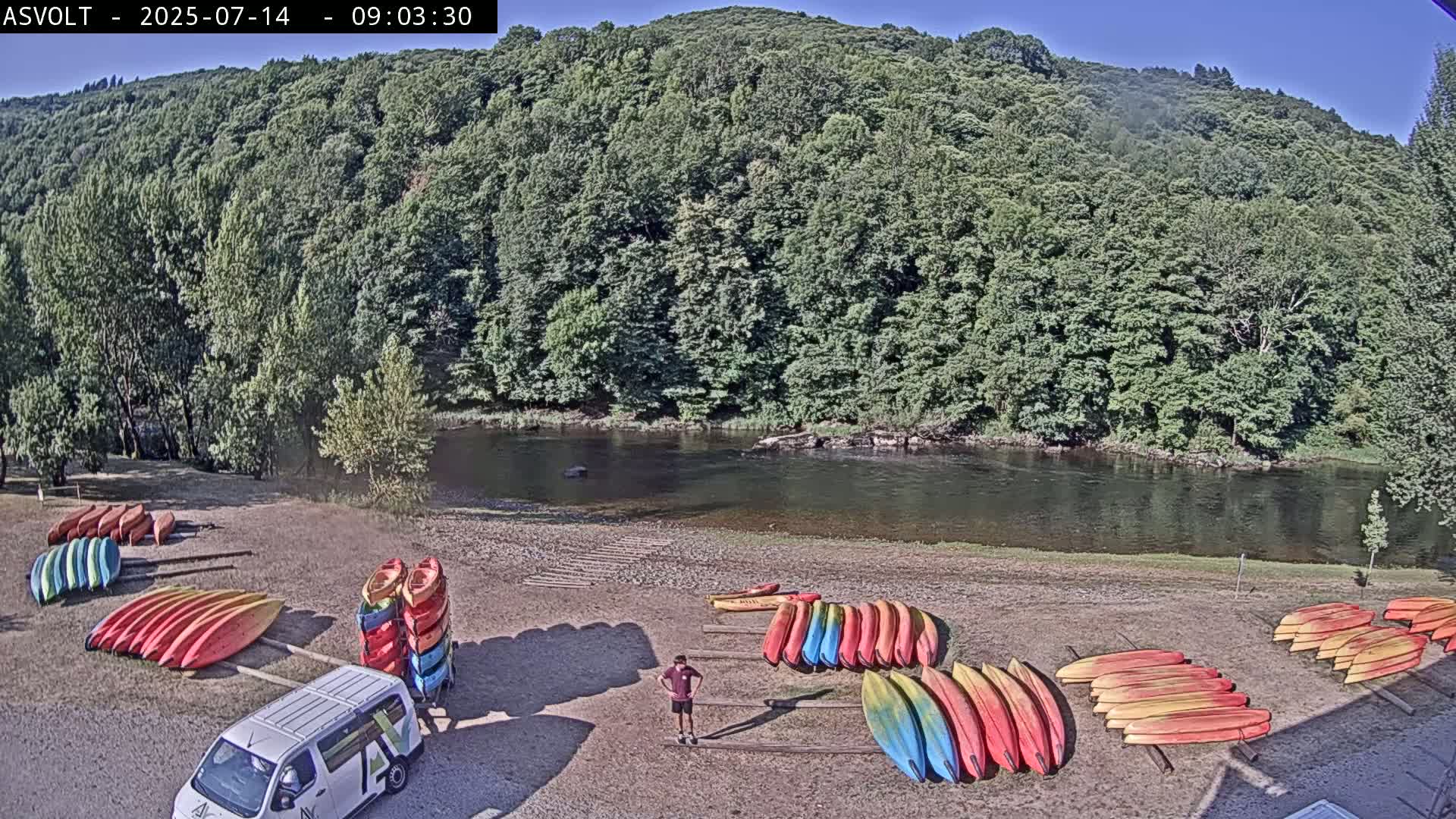 A person stands near stacks of colorful kayaks by a calm river under a clear, sunny sky, backed by a lush green hillside.