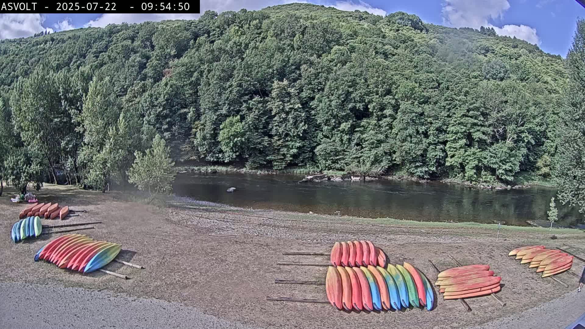 Several stacks of colorful kayaks are on a sandy shore next to a calm river, backed by a lush green forest under a partly cloudy sky.