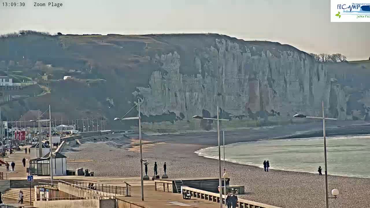 A pebble beach stretches out to a calm sea under a bright, sunny sky, with a towering cliff face rising in the background.