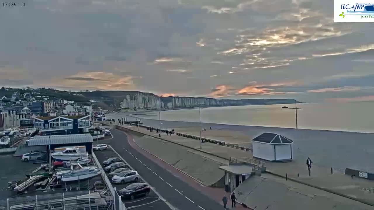 A coastal town with a harbor, beach, and white cliffs is seen at sunset under a cloudy sky.