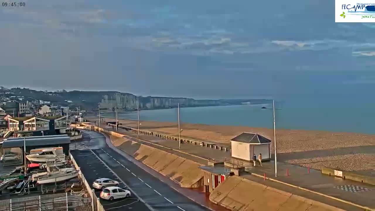 A mostly sunny day shows a beach with a calm sea, white cliffs in the distance, and a road leading to a harbor with parked cars and boats.
