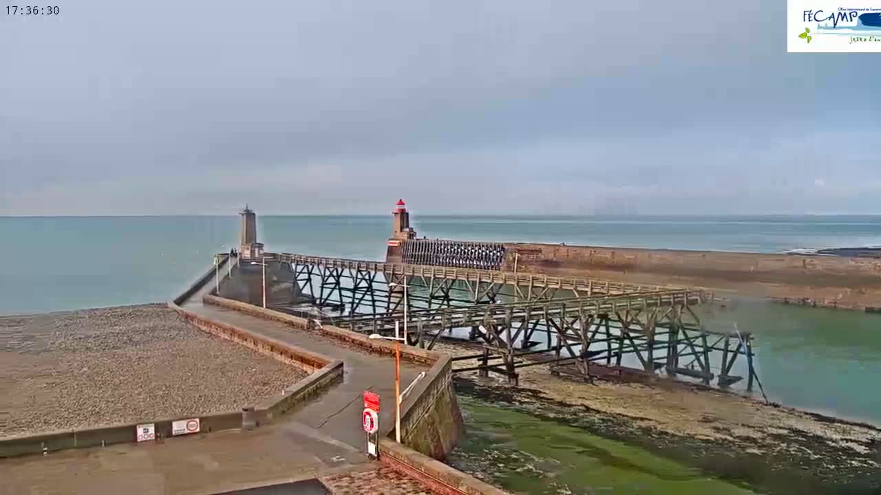 A long wooden pier with a lighthouse extends into a calm, light-blue ocean under an overcast sky.