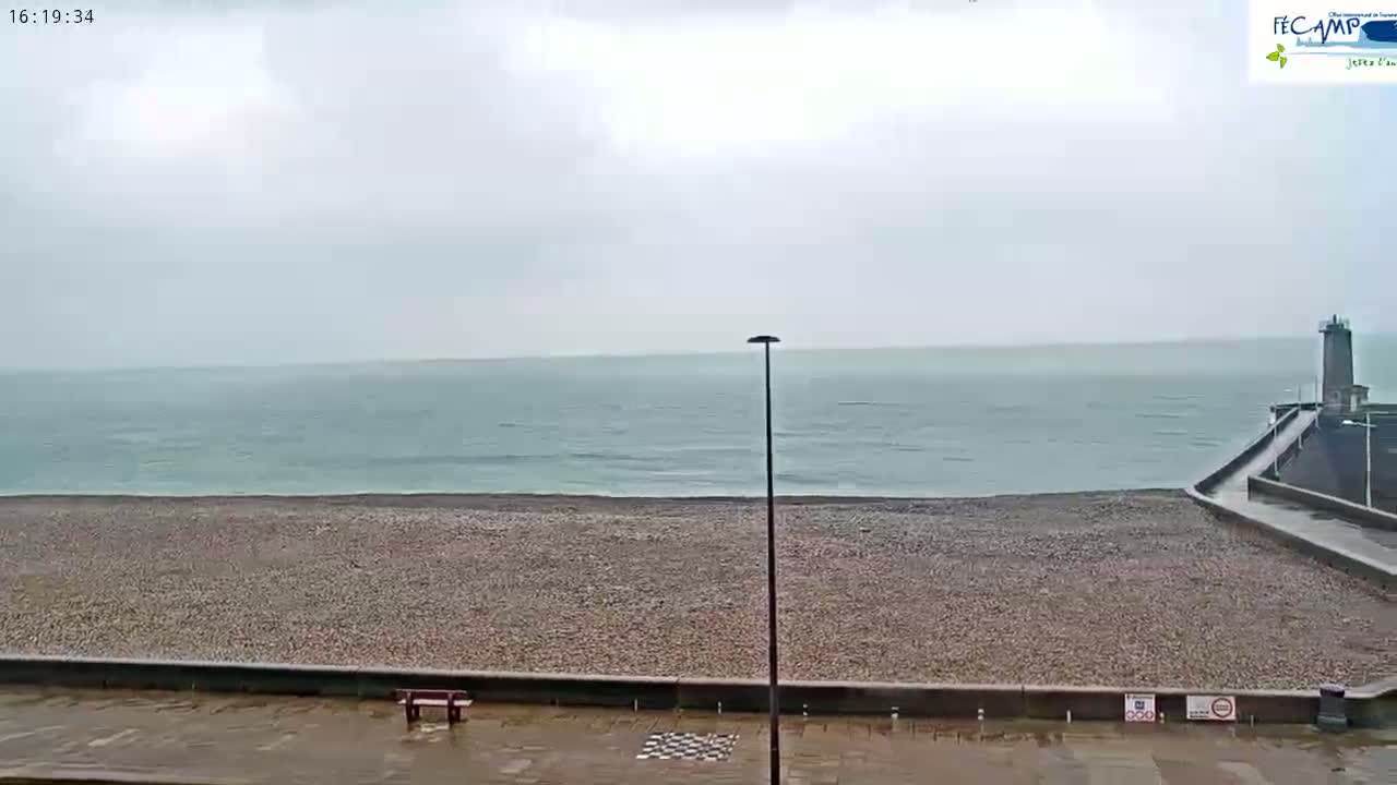 A pebble beach stretches to a calm, grey sea under an overcast sky, with a lighthouse visible in the distance to the right.
