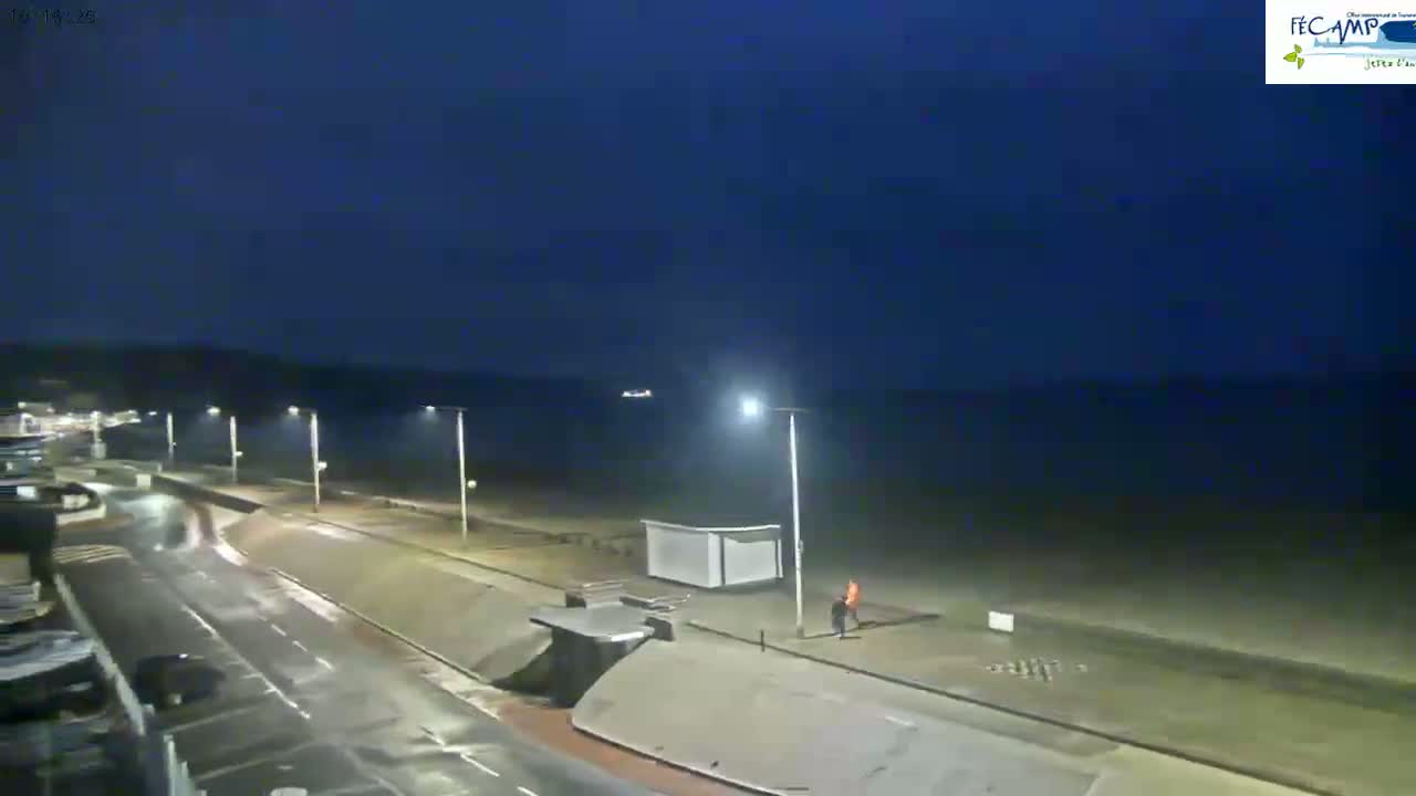 A nighttime view of a beachside road with a few people walking near a small structure, under a dark, clear sky.