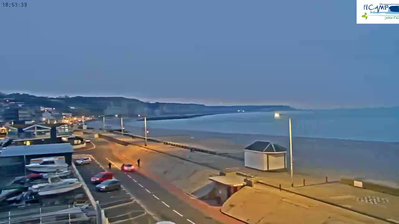An empty beach at dusk is seen from above, with a road and buildings in the foreground and calm water in the background.