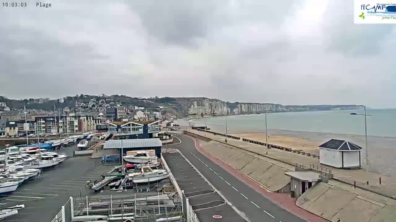 A coastal town with a marina and white cliffs is visible under an overcast sky, with a calm sea stretching out to the horizon.