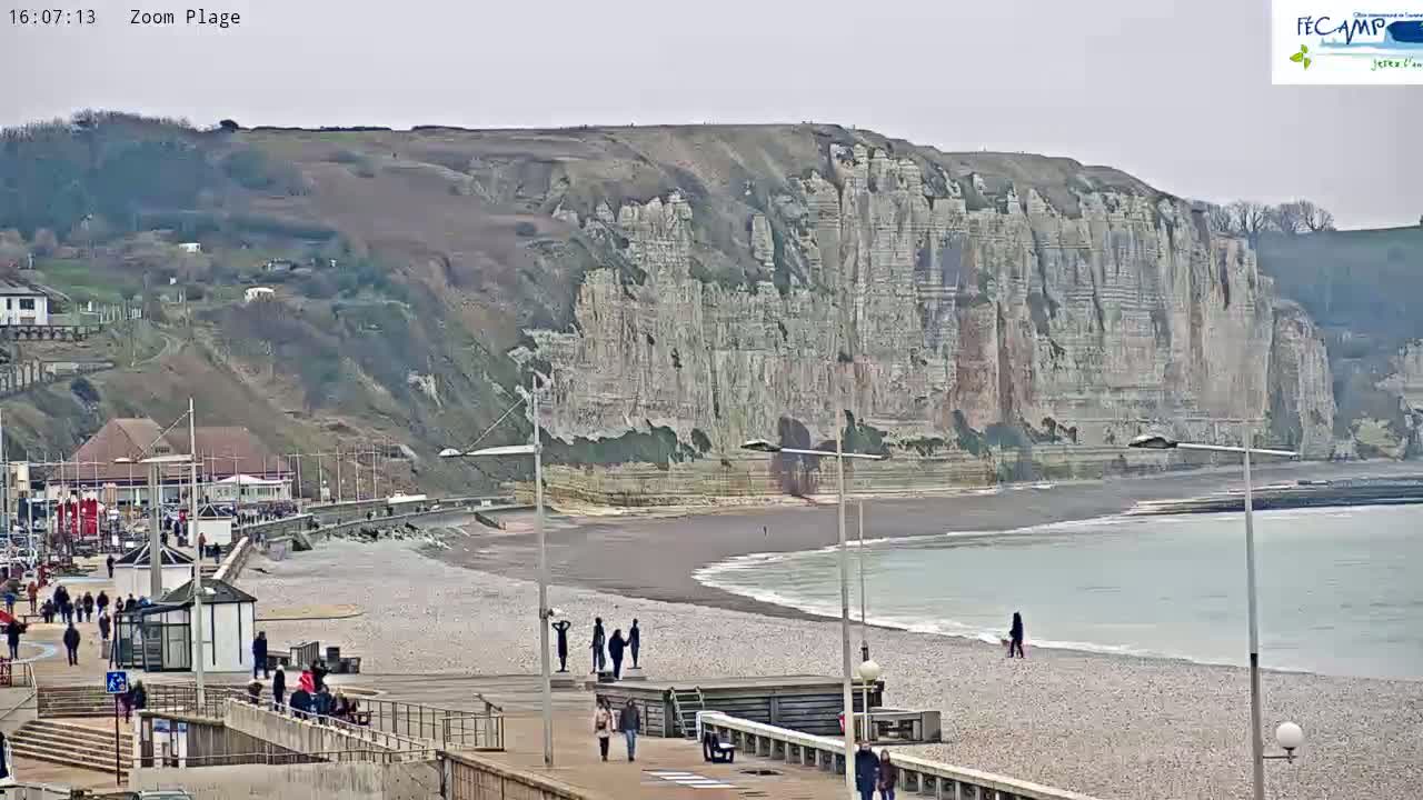 A pebble beach with people walking along a promenade, overlooking a large, white cliff face under an overcast sky.