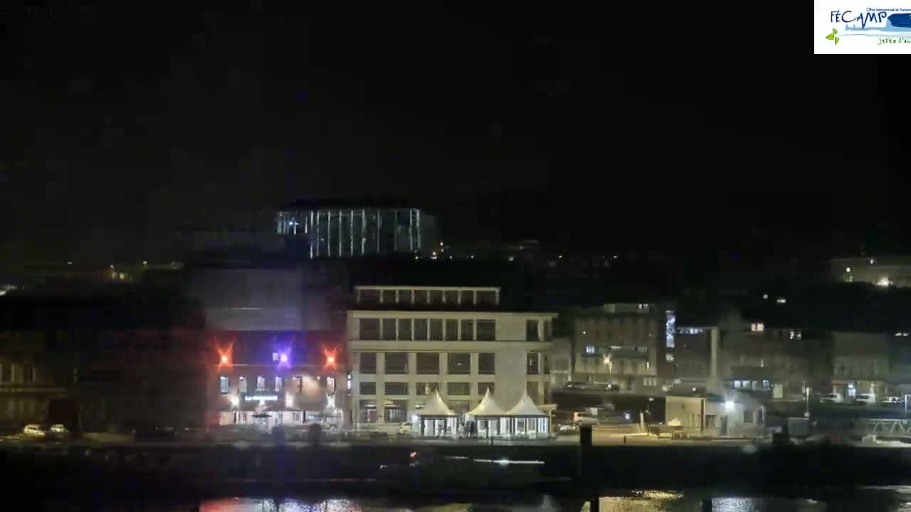 A nighttime view of a waterfront city with several buildings illuminated, some temporary tents near the water's edge, and a calm body of water in the foreground.