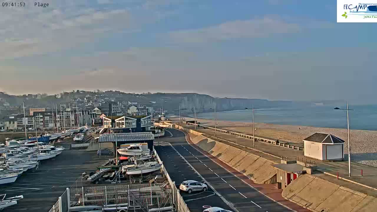 A harbor filled with boats is next to a road that runs along a beach and high, white cliffs under a partly cloudy sky.