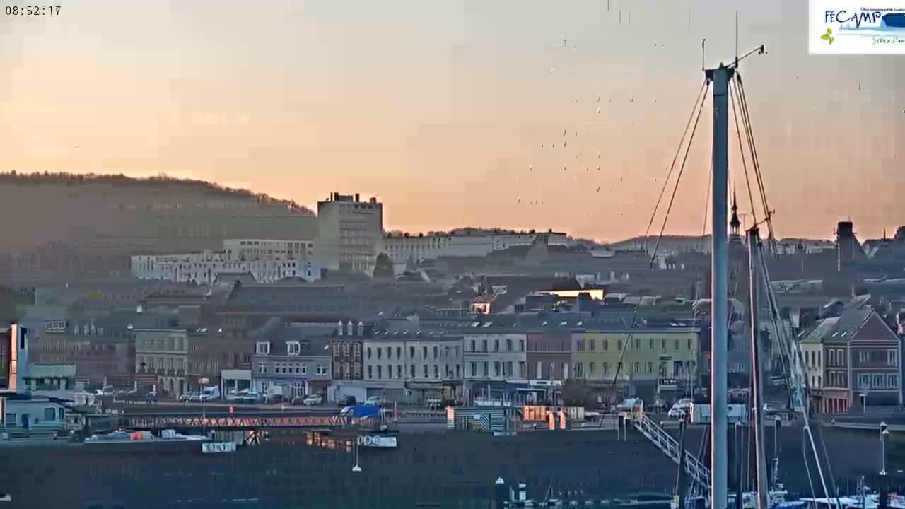 A harbor town at sunset is shown, with numerous buildings and a sailboat mast in the foreground, and a flock of birds flying above.