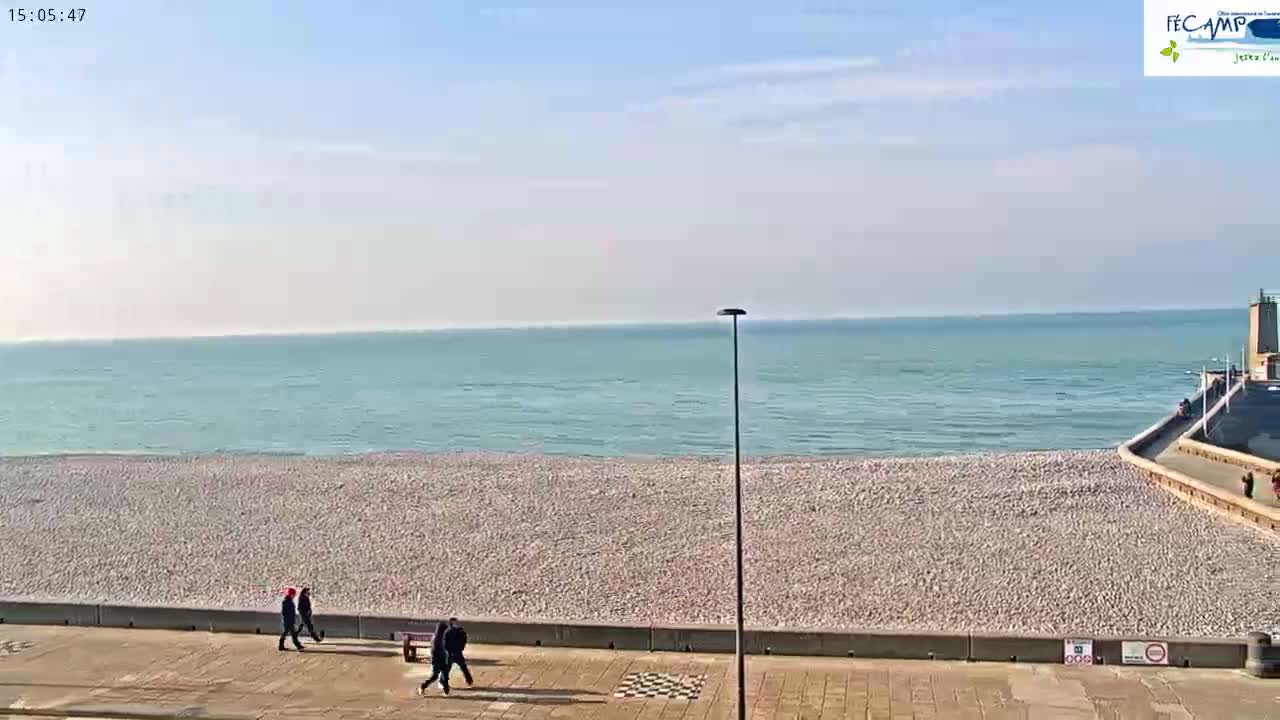 A pebble beach stretches along a calm, light-blue ocean under a mostly clear, bright sky, with a few people walking along the promenade.