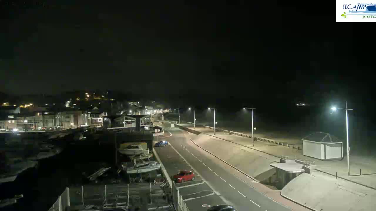 A nighttime, high-angle view shows a mostly empty road and promenade next to a beach, with buildings and boats visible in the background.