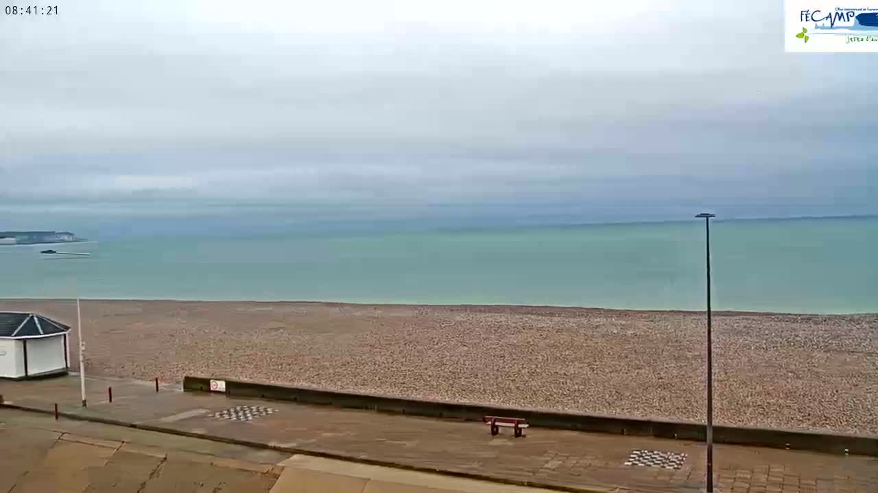 A wide shot shows a pebble beach and calm, light-green sea under an overcast sky, with a small white building and a bench visible on the promenade.