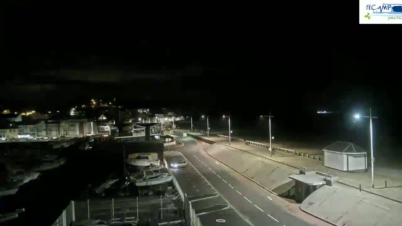 A nighttime view of a coastal road with a few cars, leading to a dark beach with some buildings and lights.