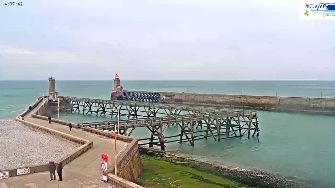 A long wooden pier extends from a rocky shoreline into a calm, teal-colored sea, with two lighthouses visible in the distance under an overcast sky.