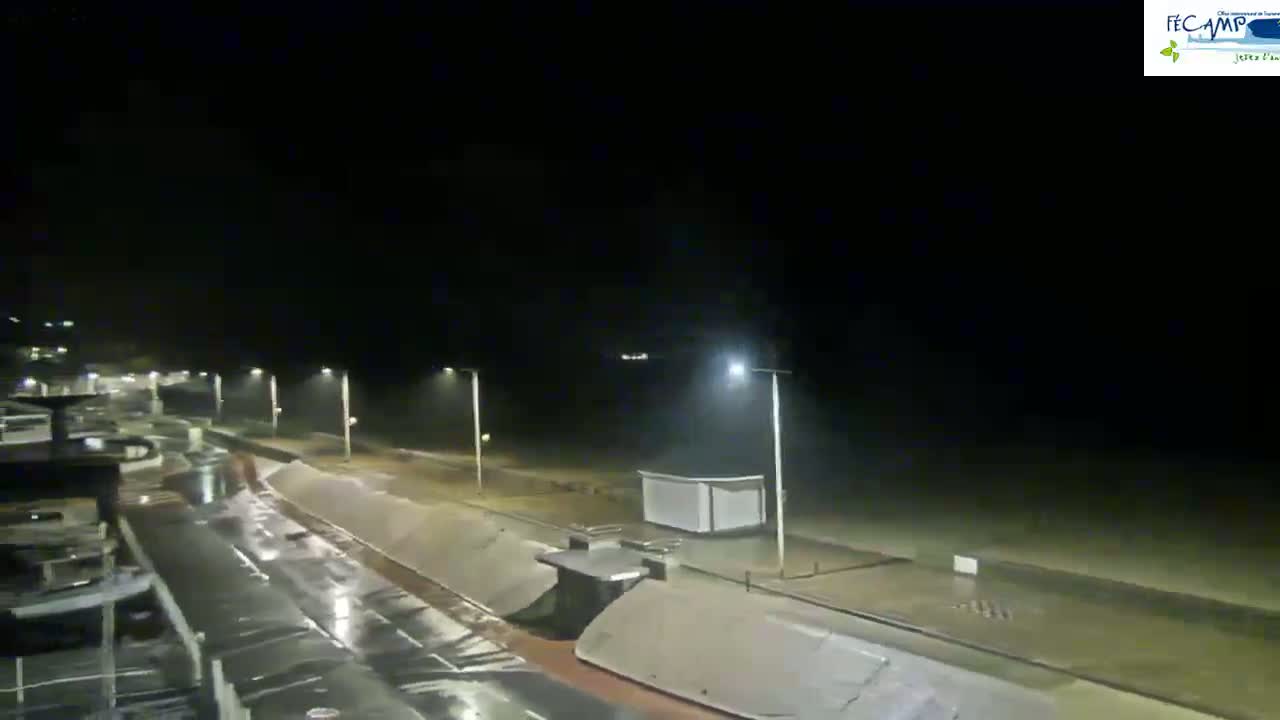 A rain-swept nighttime coastal scene shows a dark beach and promenade illuminated by streetlights, with boats visible in the foreground.