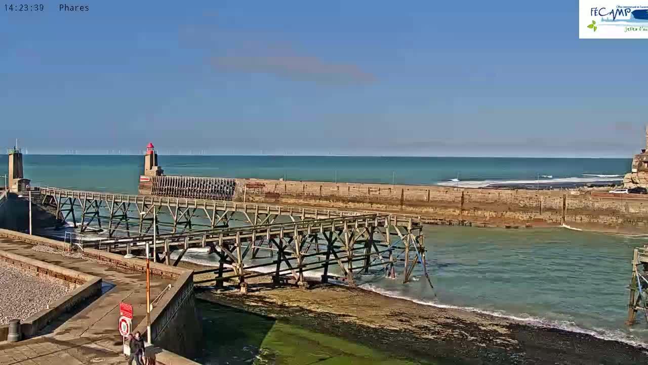 A wooden pier extends over a calm, teal-colored ocean under a clear blue sky, with a lighthouse and other harbor structures visible in the background.