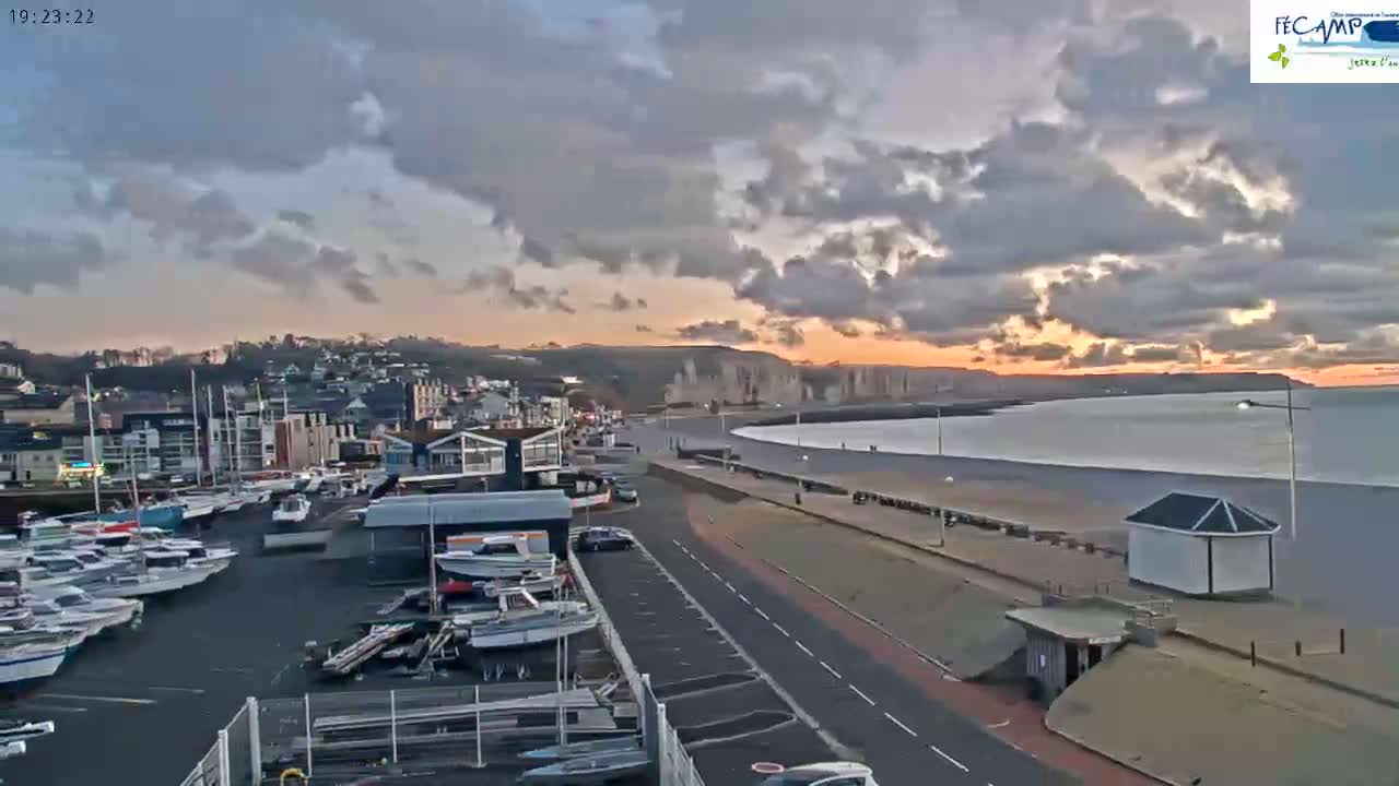 A coastal town at sunset under a partly cloudy sky, showing a harbor full of boats, a long beach, and a road leading along the waterfront.