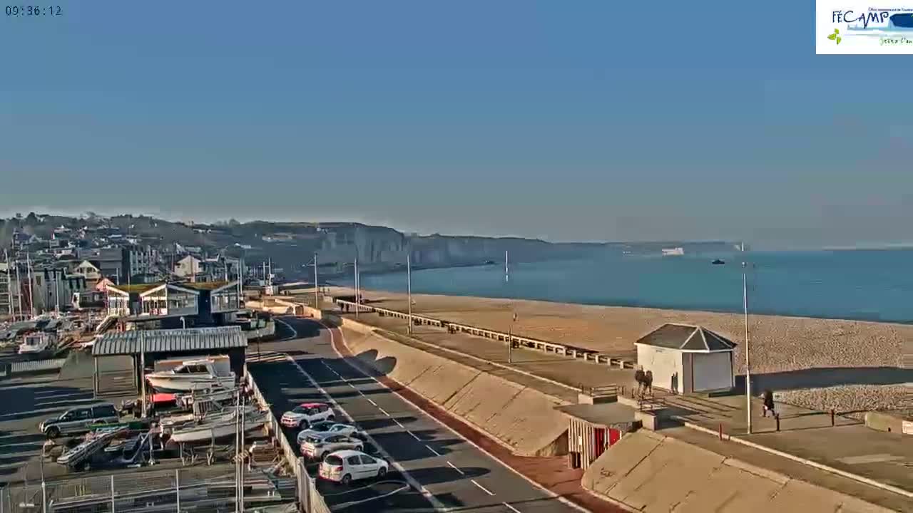 A coastal town scene on a sunny day shows a beach, calm ocean, white cliffs in the distance, parked cars, a harbor with boats, and a roadway leading towards the water.