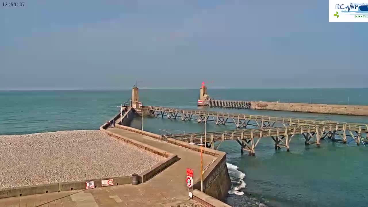 A long, stone pier extends into a calm, blue ocean, featuring two smaller piers connected by weathered wooden walkways and topped with lighthouses.
