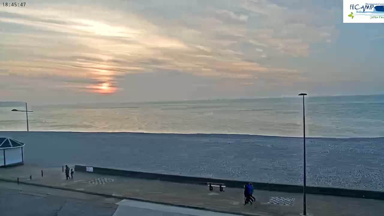A beach scene at sunset shows a calm ocean, a wide sandy beach, and a few people walking along a paved path.