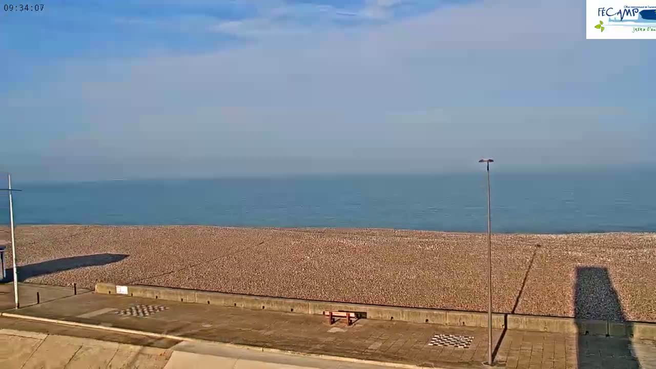 A wide, pebble beach stretches to a calm, blue ocean under a clear, bright sky.