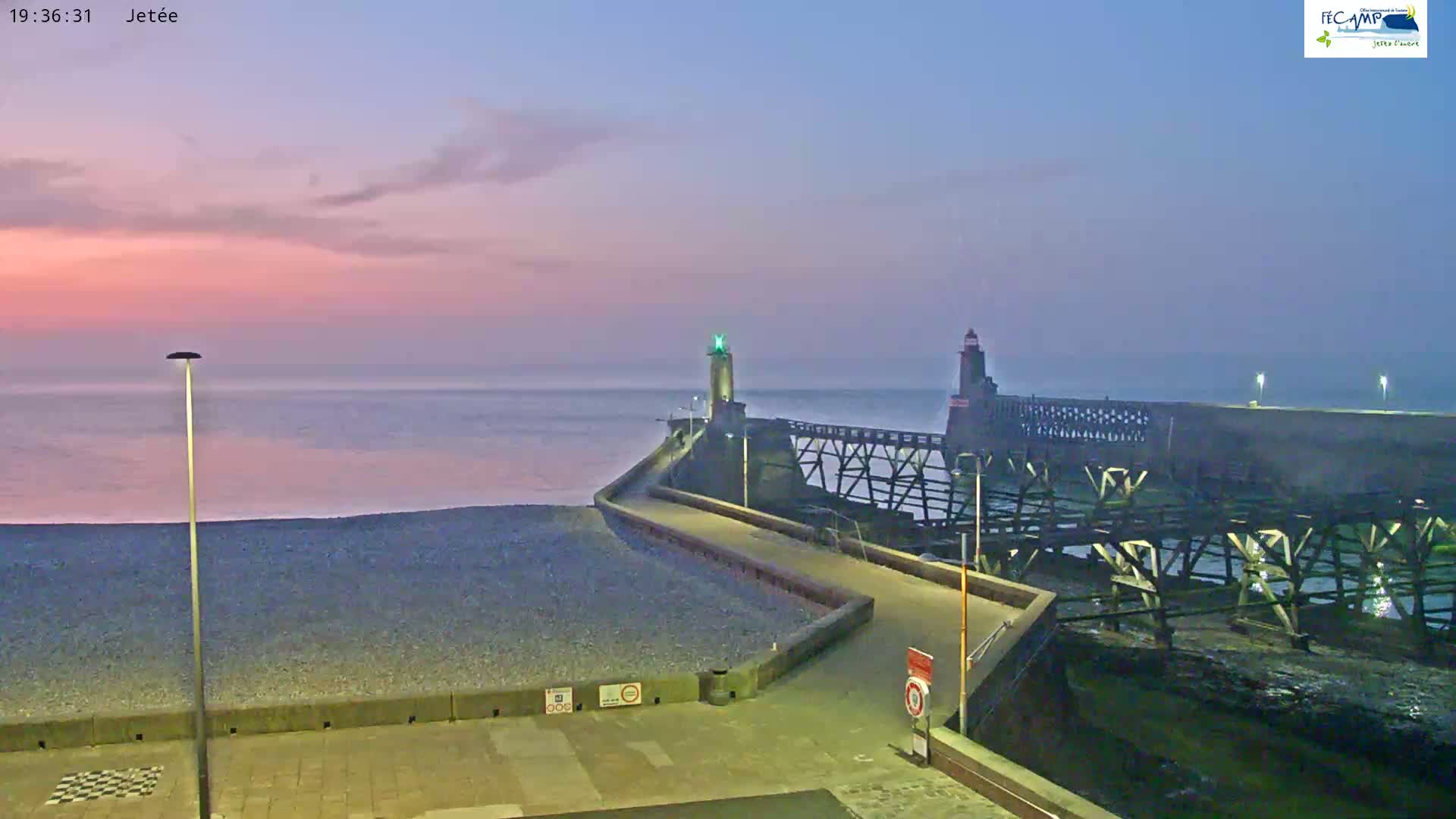 A pebble beach at twilight, with a long jetty extending into a calm sea, featuring two lighthouses.