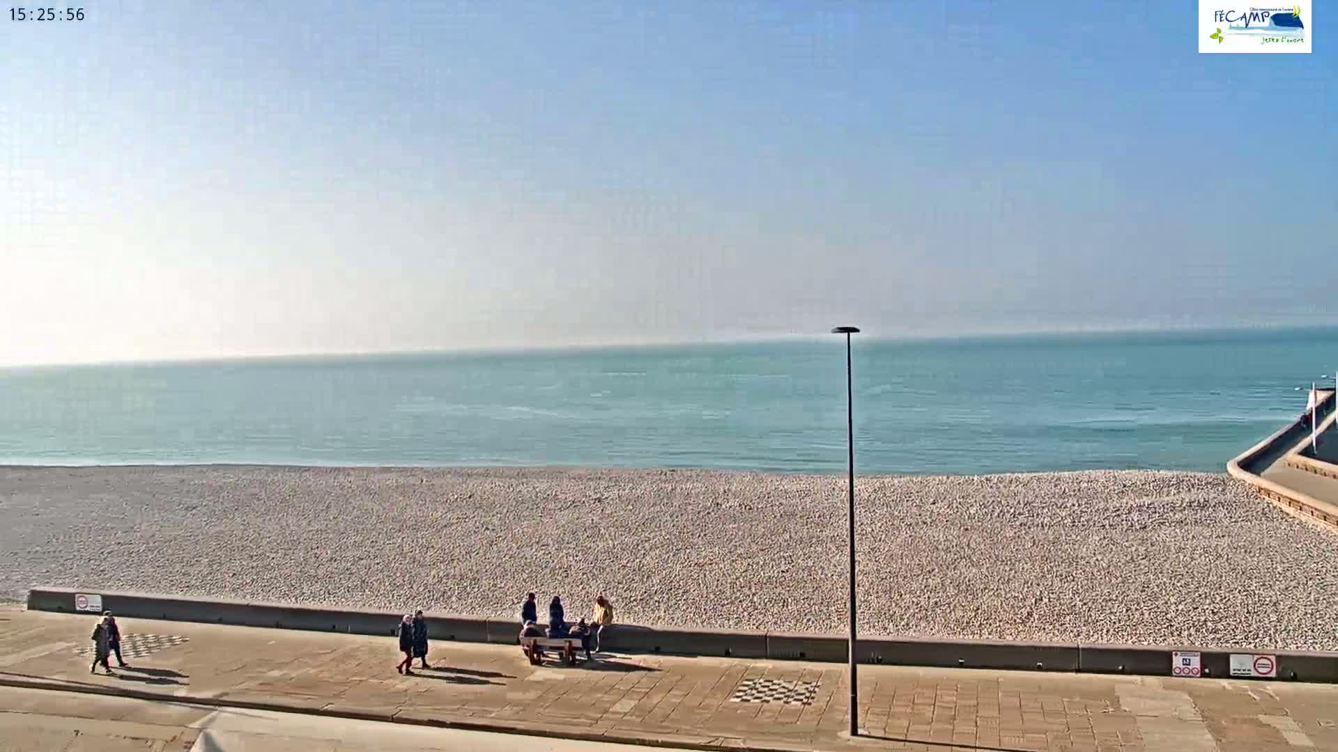 A mostly sunny day reveals a calm ocean meeting a pebble beach where several people stroll and sit on a bench.