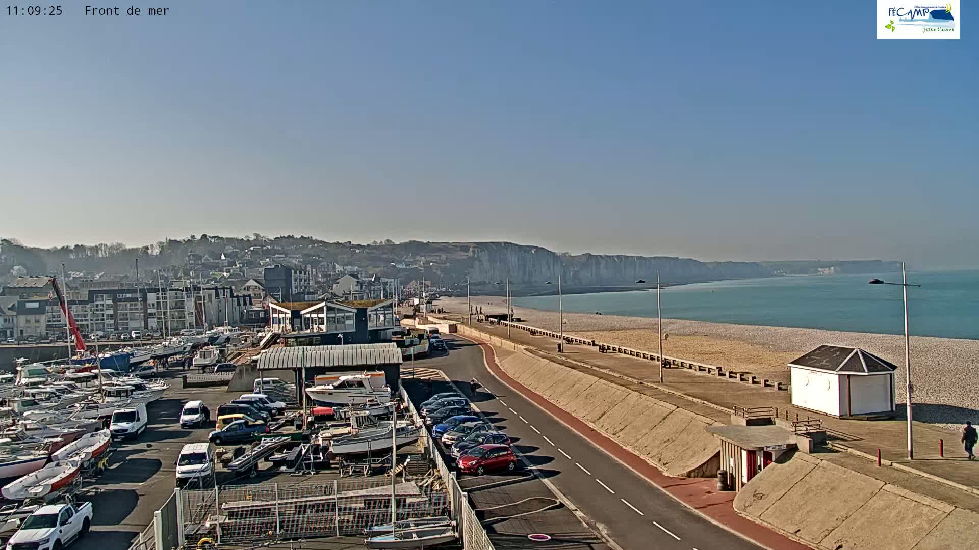 A sunny day reveals a harbor filled with boats, a paved walkway alongside a pebble beach, and towering white cliffs in the distance.