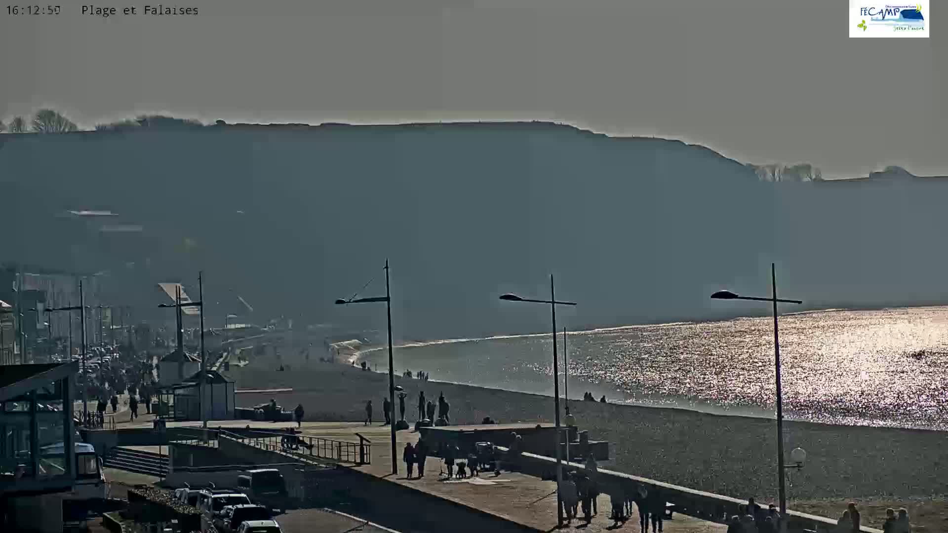 A sunny day at a beach with a pebble shoreline, a boardwalk lined with people, and large cliffs in the background.