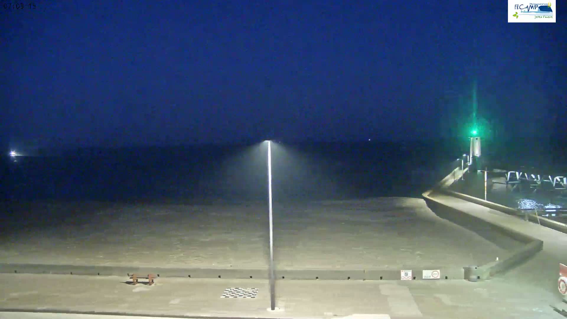 A nighttime view of a calm, dark-blue sea next to a paved walkway with a tall light pole and a small structure with a green light on the right.
