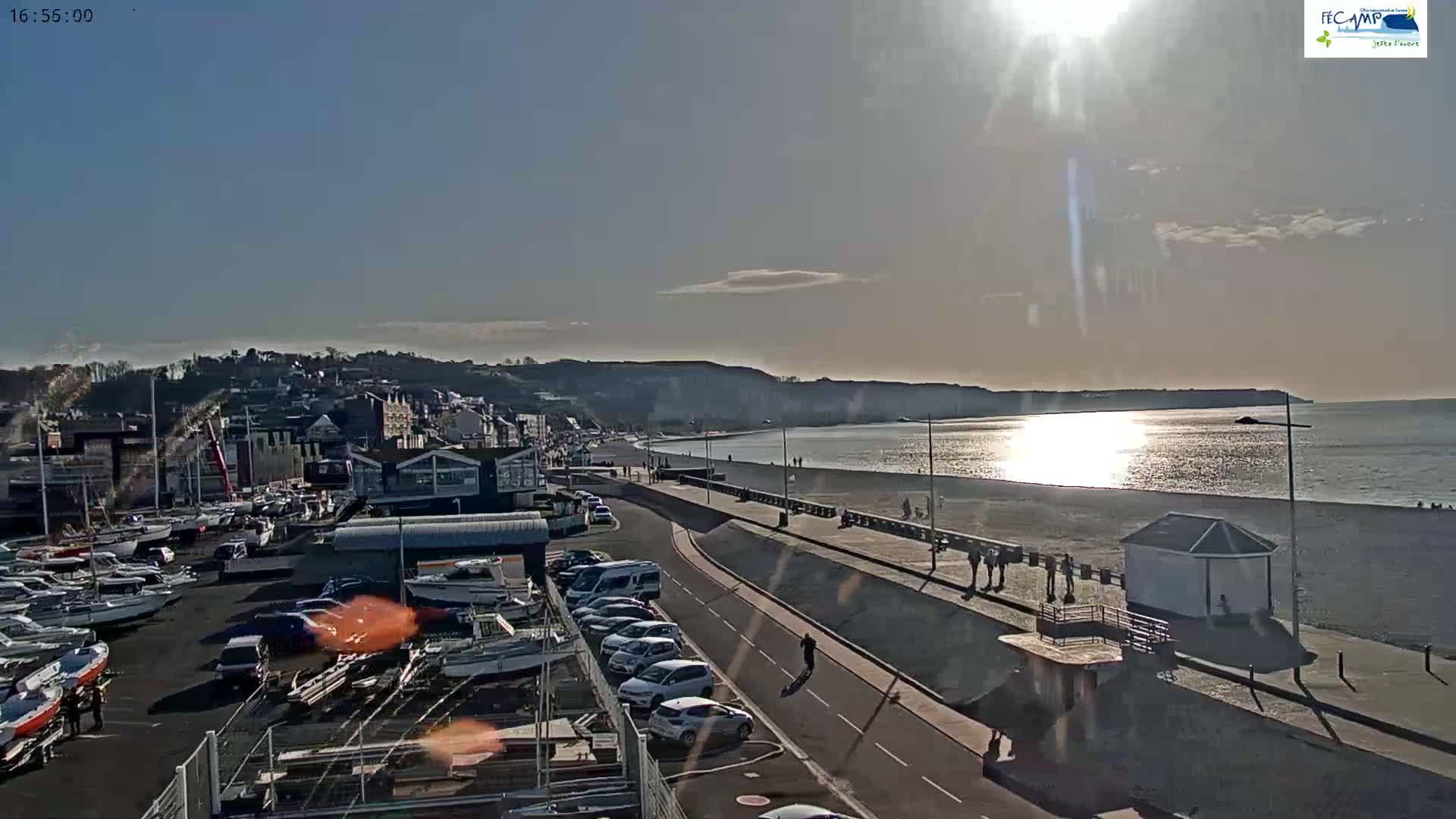 A sunny day at a seaside town shows a harbor full of boats, a road, a beach with a few people, and a shoreline with buildings and cliffs in the background.