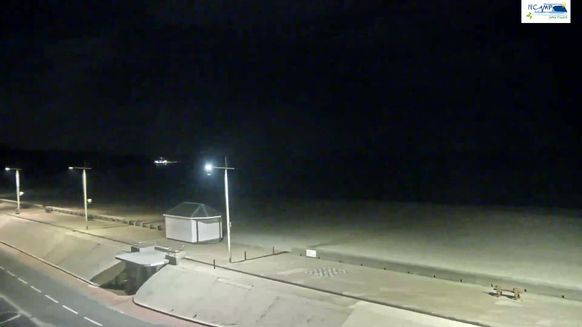 A dark, nighttime view of an empty beach with a paved walkway, streetlights, and small structures.